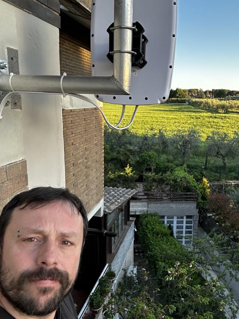 Selfie taken from below — the author at bottom-left of frame, the directional 5G antenna mounted on the building wall above him, sunlit countryside and a vineyard in the background under a clear sky