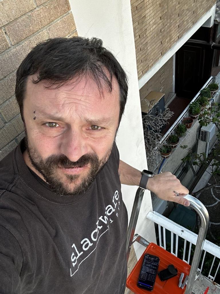 The author on a stepladder on a balcony, wearing a Slackware t-shirt, looking at the camera. A phone showing 5g-monitor signal output is propped on the ladder rail next to him; an apartment building and potted plants are visible behind